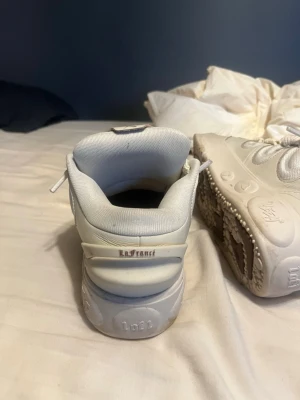 Vita La France sneakers Puma Lomeloball - Säljer ett par vita sneakers från La France med chunky sula och coola detaljer på sidan. Skorna har en modern siluett med vadderad krage och ovandel i syntetmaterial och mesh. Perfekta för dig som gillar streetwear och vill sticka ut.
