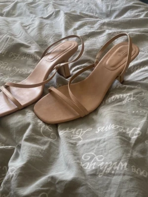 Beige sandaletter med klack - Säljer ett par stilrena beige sandaletter med smala remmar och fyrkantig tå. Skorna har en medelhög blockklack och öppen häl med enkel rem runt ankeln. Tillverkade i syntetmaterial och perfekta för dig som gillar minimalistisk design.