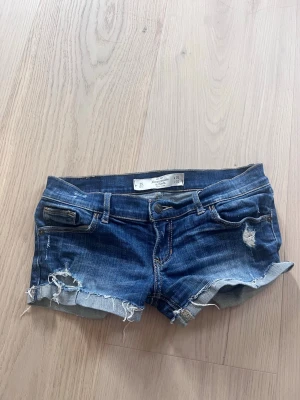 Blå vintage lågmidjade jeansshorts med slitningar - Säljer ett par blå vintage low waist lågmidjade jeansshorts från Abercrombie & fitch med råa kanter och slitna detaljer. Klassisk femficksmodell med låg midja och uppvikta benslut. Perfekta för varma dagar och en avslappnad stil. Liten repa på baksidan men inget som inte går att fixa!