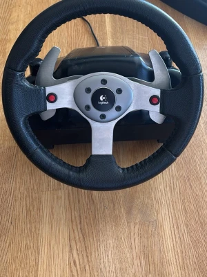 Logitech ratt till spelkonsol - Säljer en svart och silverfärgad ratt från Logitech för spelkonsol. Ratten har en robust metallram, två röda knappar och är klädd i konstläder för bra grepp. Perfekt för racing- och bilspel för en mer realistisk spelupplevelse.