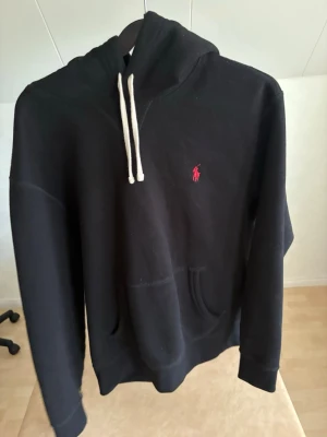Svart hoodie från Polo Ralph Lauren - Svart hoodie med vit dragsko och klassisk röd Polo Ralph Lauren-logga broderad på bröstet. Hoodien har känguruficka framtill och ribbade muddar vid ärmar och nederkant. Perfekt för en avslappnad och stilren look.