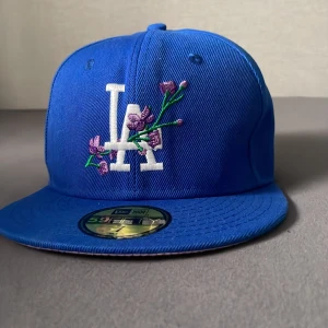 Blå LA Dodgers keps med brodyr - Snygg blå snapback-keps från New Era med LA Dodgers-logga framtill och lila blommor broderade över loggan. På sidan finns ett broderat 1988 World Series-märke. Klassisk rak skärm och justerbar passform. Perfekt för dig som vill sticka ut med stilren streetwear.