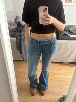 Blå bootcut lågmidjade jeans med fickor - Säljer ett par blå bootcut jeans med hål på knäna och låret. Jeansen har två stora framfickor och dubbla knappar.