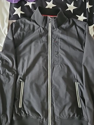 Svart Superdry vindjacka med vita dragkedjor - Snygg svart vindjacka med vita dragkedjor framtill och på fickorna. Jackan har en bröstficka med knappdetalj och elastiska muddar vid ärmslut och nederkant. Tillverkad i lätt och vindtätt material, perfekt för blåsiga dagar. Liten reva bak i nacken som syns på bild. 