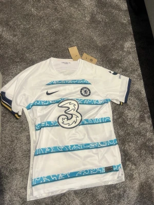 Chelsea FC bortatröja James 24 - Chelsea FC bortatröja i vitt med blå detaljer och tryck. Tröjan har blå ränder med grafiskt mönster, klubbmärke och Nike-logga på bröstet samt sponsortryck. På ryggen står det 'James 24' i stora blå siffror. Tillverkad i lätt och ventilerande material.