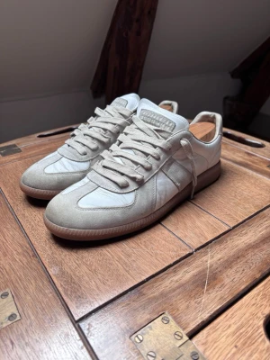 Vita sneakers från Maison Margiela - Snygga vita sneakers från Maison Margiela med beige mockadetaljer och klassisk gummisula. Skorna har låga skaft, vita skosnören och en stilren design i både läder och mocka. Perfekta för dig som gillar minimalistisk och clean stil.