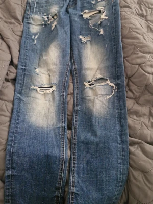 Blå slitna jeans med hål - Säljer ett par blå jeans med slitningar och stora hål på knäna och låren. Jeansen har en klassisk femficksdesign och är tillverkade i denim. De har en rak passform och tydliga blekta partier för en cool look.