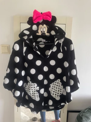 Mimmi Pigg poncho med huva - Supermysigw svart poncho i fleece med vita prickar och två fickor fram. Huvan har Mimmi Pigg-ansikte, öron och en stor rosa rosett. Perfekt för chill dagar hemma när du vill vara både bekväm och söt.