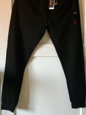 Svarta mjukisbyxor Polo Ralph Lauren - Svarta mjukisbyxor från Polo Ralph Lauren med röd logga broderad på vänster ben. Byxorna har dragsko i midjan, muddar vid bensluten och sidofickor. Tillverkade i mjukt material som känns skönt mot huden. Perfekta för en chill och avslappnad stil.