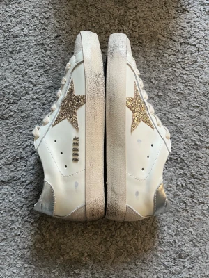 Golden Goose Superstar sneakers glitter - Snygga vita Golden Goose Superstar sneakers med glitterstjärna på sidan och silverfärgad häl. Skorna har beige snörning och slitna detaljer på sulan för en cool vintage-look. Tillverkade i läder med textildetaljer och GGDB-logga på sidan.