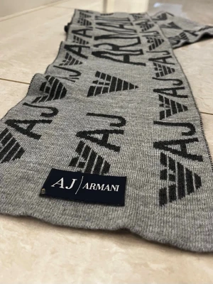 Grå halsduk från Armani Jeans - Snygg grå halsduk från Armani Jeans med svart logotyp och textmönster över hela halsduken. Tillverkad i mjukt stickat material som känns skönt mot huden. Perfekt accessoar för att lyfta din outfit med en exklusiv touch.