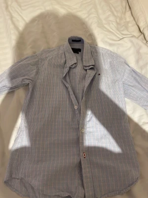 Rutig skjorta från Tommy Hilfiger - Snygg rutig skjorta från Tommy Hilfiger i ljusblått och vitt med diskret röd detalj. Skjortan har klassisk krage, knappar framtill och lång ärm. Tillverkad i bomull för en skön känsla och slim fit-passform.