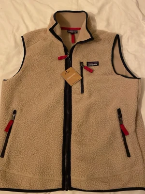 Beige fleeceväst från Patagonia - Säljer en beige fleeceväst från Patagonia med svart kantband och röda dragkedjor. Västen har hög krage, två fickor med dragkedja och en bröstficka. Tillverkad i mjuk fleece som håller värmen och har en sportig look.
