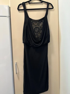 Svart klänning med draperad front - Svart kortklänning med breda axelband och draperad front. Framsidan har ett dekorativt broderi med pärlor i guldtoner. Klänningen har en lösare passform upptill och är ärmlös, perfekt för dig som gillar en unik och elegant look.OBS: FRAKTAR INOM 24H!🩷