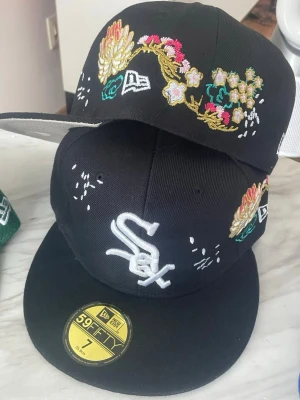 Svart New Era 59FIFTY White Sox keps - Säljer en svart fitted keps från New Era 59FIFTY med White Sox-logga framtill. Kepsen har broderade blommor i gult, rosa och grönt på sidorna samt vita detaljer. Klassisk rak skärm och broderade ventilationshål. Storlek 7 (55,8 cm).