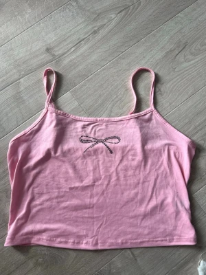 Rosa linnetopp med rosett från SHEIN - Söt rosa linnetopp från SHEIN med tunna axelband och en liten rosett i strass framtill. Toppen är croppad och gjord i mjuk bomull, perfekt för en trendig och avslappnad stil.