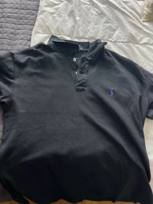 Svart pikétröja från Polo Ralph Lauren - Klassisk svart pikétröja från Polo Ralph Lauren med lila broderad logga på bröstet. Tröjan har korta ärmar, krage och knappar framtill. Tillverkad i 100% bomull för en skön och avslappnad känsla.