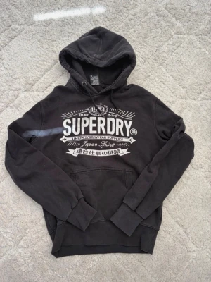 Svart hoodie från Superdry - Säljer en svart hoodie från Superdry med stor vit logga och tryck på bröstet. Hoodien har huva med dragsko och en klassisk magficka. Tillverkad i mjukt bomullsmaterial 