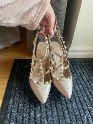 Beiga klackar med nitar och remmar - Snygga beige pumps med spetsig tå och flera remmar dekorerade med guldfärgade nitar. Skorna har en lackad finish och justerbara spännen vid ankeln. Perfekta för dig som vill ha en edgy men elegant look.