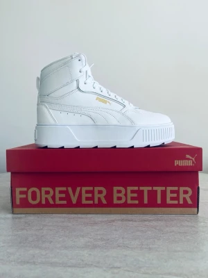 Vita höga sneakers från Puma - Stilrena vita sneakers från Puma med hög siluett och chunky sula. Skorna har detaljer i guld, klassisk snörning och är tillverkade i slätt läder. Perfekta för dig som vill ha en clean och modern look med streetkänsla.