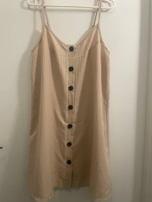 Beige sommarklänning med knappar - Snygg beige klänning med tunna axelband och svarta knappar framtill. Klänningen är kort och har en rak passform, perfekt för varma dagar. Tillverkad i ett lätt och luftigt material som känns skönt mot huden.