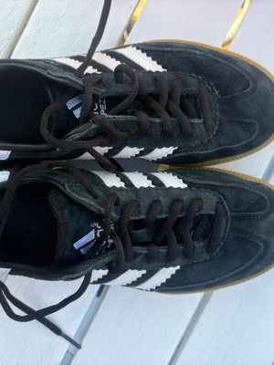 Adidas svartvita sneakers Samba - Klassiska Adidas Samba sneakers i svart mocka med vita läderdetaljer och bruna gummisulor. Skorna har svarta skosnören och den ikoniska tre ränder-designen på sidorna. Perfekta för en sportig och avslappnad stil.