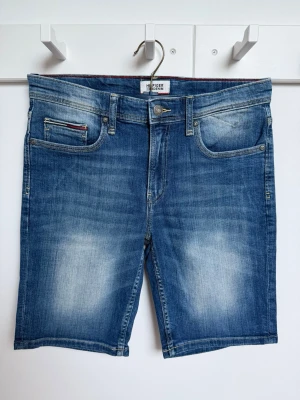 Blå denimshorts från Hilfiger Denim - Snygga blå jeansshorts från Hilfiger Denim med klassisk femficksdesign och subtila slitningar. Shortsens midja är normal och de har en knapp- och dragkedjeförslutning. Perfekta för en avslappnad och cool look. Storlek 176/S. Mycket bra skick ser ut som nya.