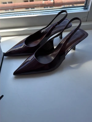 Vinröda lackade slingback-pumps - Säljer ett par vinröda slingback-pumps. Skorna har spetsig tå, lackad finish och smal klack. Tillverkade i glansigt syntetmaterial med öppen häl och elegant design. Perfekta för dig som vill ha en stilren och trendig look.