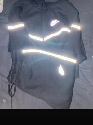 Nike tech reflective - Snygg svart hoodie från Nike med dragkedja och huva. Hoodien har vita reflexdetaljer över bröstet, armarna och vid fickorna samt Nike-logga på bröstet och ärmen. Tillverkad i mjukt material som känns skönt mot huden. Perfekt för dig som vill sticka ut lite extra.