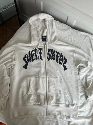 Vit zip hoodie SWEET SKTBS - Vit hoodie med dragkedja från SWEET SKTBS. Stor svart text i grafisk stil över bröstet. Klassisk känguruficka framtill och ribbade muddar. Tillverkad i mjuk bomullsmix och har en stor huva för extra chill vibe.