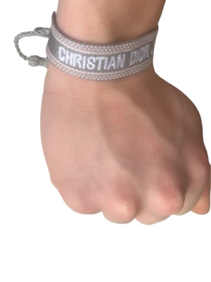 Grått Christian Dior tygarmband - Snyggt grått tygarmband från Christian Dior med texten 'CHRISTIAN DIOR J'ADIOR' broderad i vitt. Armbandet har justerbar knytning och en stilren, modern look som passar perfekt till din outfit.