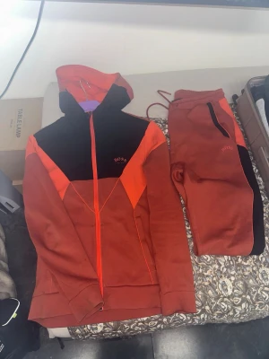 Röd-orange mjukisdress från BOSS - Säljer ett par snygga mjukisbyxor från BOSS i röd-orange färg med svarta detaljer. Byxorna har resår i midjan och dragkedjeficka. Matchande hoodie med dragkedja och huva. Perfekt för chill dagar eller när du vill vara bekväm men ändå se fresh ut.