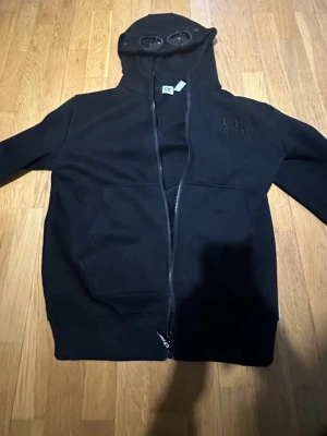 Svart hoodie med goggles från C.P. Company - Svart hoodie från C.P. Company med hel dragkedja och ikoniska goggles i huvan. Tröjan har broderad logga på bröstet och stora fickor framtill. Tillverkad i mjukt material som känns skönt mot huden. Perfekt för dig som gillar streetwear och unika detaljer. Dragkedjan är lite trög, därav priset. Går att förhandla.   Dm vid minsta fråga! 