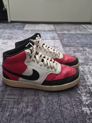 Nike court Visions red - Säljer ett par snygga Nike sneakers med högt skaft. Skorna har en vit bas med röda och svarta detaljer, klassisk svart swoosh på sidan och vita skosnören. Ovandelen är i läder och perforerad framtill för extra ventilation.
