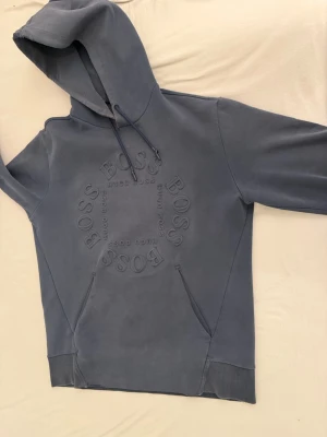 Blå hoodie från Hugo Boss - Snygg blå hoodie från Hugo Boss med stor logga i relief på bröstet. Hoodien har huva med dragsko, känguruficka och ribbade muddar vid ärmslut och nederkant. Perfekt för en avslappnad och trendig stil.