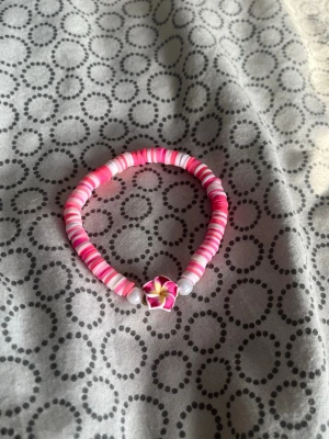 Rosa pärlarmband med blomdetalj - Sött armband med platta pärlor i olika rosa och vita nyanser. I mitten sitter en dekorativ blomma i rosa, vitt och gult samt två vita pärlor på varsin sida om blomman. Perfekt accessoar för dig som gillar färg och unika detaljer.