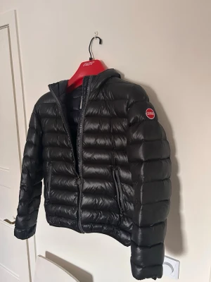 Svart pufferjacka från Colmar - Snygg svart pufferjacka från Colmar med huva och dragkedja framtill. Jackan har två sidofickor med dragkedja och ett Colmar-märke på vänster ärm. Tillverkad i glansigt material som ger en modern look och håller dig varm under kalla dagar.