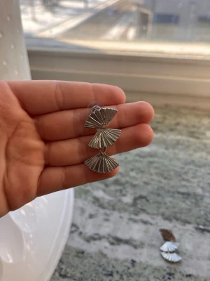 Silvriga solfjäderformade örhängen - Unika örhängen i silverfärgad metall med tre solfjäderformade delar som hänger i rad. De har en modern och trendig design med räfflade detaljer som fångar ljuset snyggt. Perfekta för dig som vill sticka ut med accessoarerna.