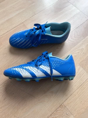 Blå Adidas Predator fotbollsskor - Snygga blå Adidas Predator fotbollsskor med vita detaljer och klassiska ränder på sidan. Skorna har snörning och är tillverkade i syntetmaterial med dobbar för bra grepp på planen. Perfekta för dig som vill sticka ut på fotbollsplanen.