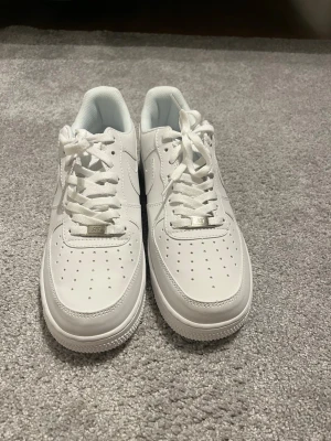 Nike Air Force 1 vita sneakers(minst 400kr) - Klassiska helvita Nike Air Force 1 sneakers i lågt utförande. Skorna har ovandel i slätt läder, perforeringar på tån för ventilation och vit sula med AIR-detalj. Swoosh-logga på sidan och vita skosnören. Perfekta för en clean och stilren look.