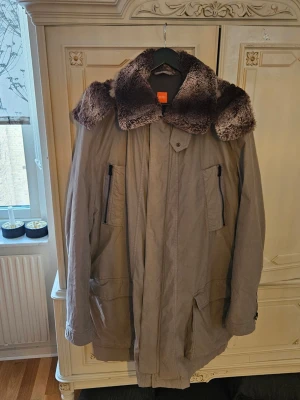Beige parkas med fuskpäls - Stilren beige parkas från Hugo Boss Orange med stor avtagbar huva. Och bred krage i mörkbrun fuskpäls. Jackan har flera praktiska fickor med dragkedja och knappstängning framtill. Perfekt för kalla dagar och ger en cool, avslappnad look. Jackan kan även bli en vårjacka då innefodret är avtagbart som luvan.