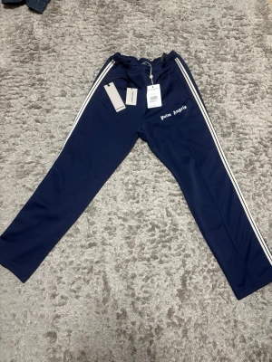 Palm angels byxor - Säljer ett par mörkblåa mjukisbyxor från Palm Angels med vita sidoränder och logga tryckt på vänster ben. Byxorna har resår i midjan och raka ben. Perfekta för en chill och trendig look. Köpta på vestiaire så har äkthetsverifickation på dem. Äldre modell. Skriv vid minska fråga.