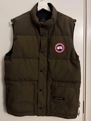 Olivgrön dunväst från Canada Goose - Säljer en olivgrön dunväst från Canada Goose med klassisk logga på bröstet och två stora fickor framtill. Västen har hög krage, tryckknappar och är fodrad med dun för extra värme. Perfekt för lager-på-lager och streetwear-stil. OBS! Kvittot finns inte kvar! 