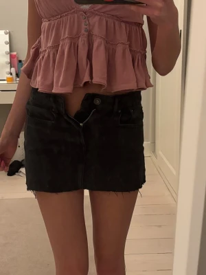 Svart jeanskjol med rå kant - Säljer en svart kort jeanskjol med rå, fransig kant nertill och klassisk femficksdesign. Kjolen har knapp och dragkedja framtill och är perfekt för en avslappnad och trendig look.