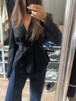 Svart kavaj med bälte och markerad midja - Snygg svart kavaj med bälte i midjan som ger en markerad siluett. Kavajen har breda slag, långa ärmar och knäppning framtill. Passar perfekt till jeans eller kostymbyxor för en trendig look. Materialet ser ut att vara bomull eller en bomullsblandning. Står att de är storlek L men skulle säga att den passar allt från Xs-M😇