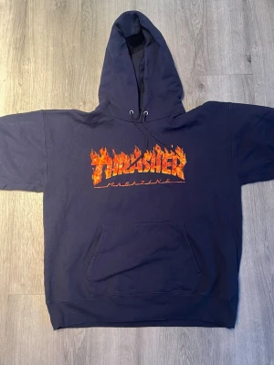 Mörkblå Thrasher hoodie - Snygg mörkblå hoodie från Thrasher med det klassiska flames-trycket i orange och gult på bröstet. Hoodien har stor huva, magficka och ribbade muddar. Perfekt för dig som gillar streetwear och skatekultur.