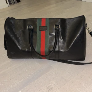 Gucci svart duffelbag med röd/grön rand - Snygg svart weekendbag från Gucci med klassisk röd och grön rand i mitten. Väskan har dubbla handtag, avtagbar axelrem och diskret GG-mönster i lädret. Perfekt för resor eller gymmet när du vill ha en stilren och lyxig look.