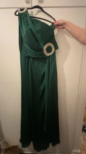Grön långklänning med pärldetaljer - Elegant grön långklänning med en axel och flödande ärm. Klänningen har två stora pärlprydda ringar, en vid axeln och en i midjan, som ger en lyxig känsla. Materialet är mjukt och faller snyggt. Perfekt för dig som vill sticka ut på bal eller fest. Köpt i Skopje så vet inte var den är köpt för lapp bortklippt.  Storlek annorlunda men den är ungefär 38/40 i vanlig storlek. SAKNAS EN PALJETT.  
