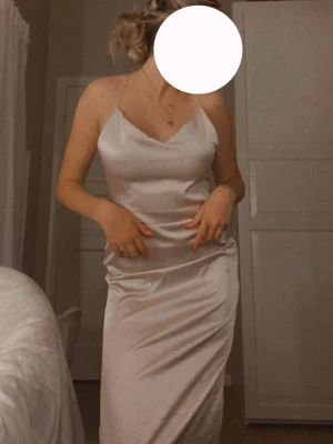 Ljus klänning med snörning - Elegant ljus klänning i sidenliknande material med tunna axelband och öppen rygg med snörning. Klänningen har en slits på sidan, klänningen går inte ända ner till fötterna. Endast använd en gång.