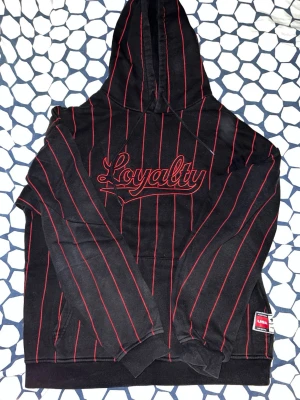 Svart hoodie med röda ränder Loyalty - Snygg svart hoodie från Loyalty med tunna röda vertikala ränder och broderad text 'Loyalty' på bröstet. Klassisk huva och känguruficka framtill. Ribbstickade muddar vid ärmslut och nederkant. Märkeslapp nertill. Perfekt för en streetwear-look.
